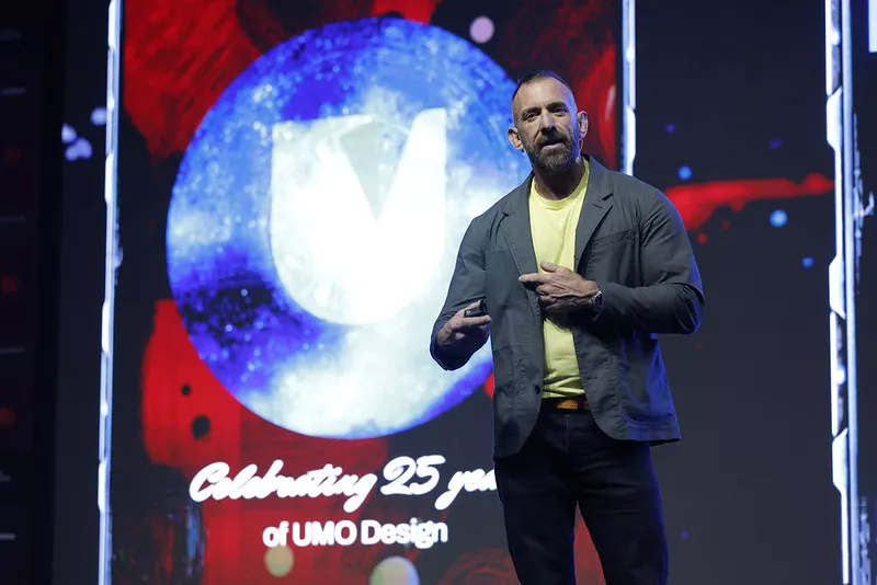 A speaker presenting on stage at a design conference with a large illuminated screen behind them